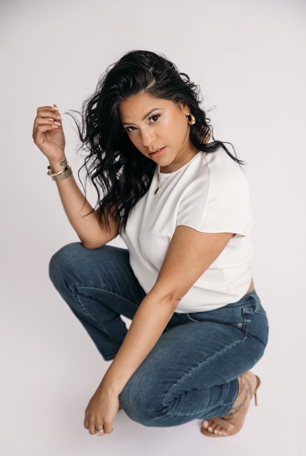Shay in a confident crouched studio portrait wearing a white top and jeans.