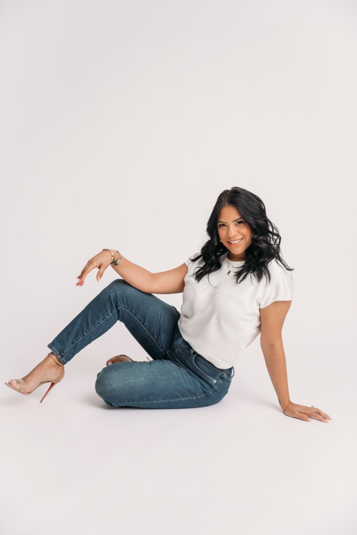 Shay smiling in a seated studio portrait against a soft neutral background.